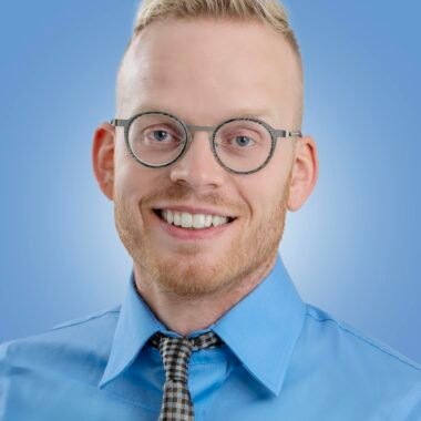 A confident and smiling man in a blue shirt and glasses against a plain background, showcasing a friendly demeanor.