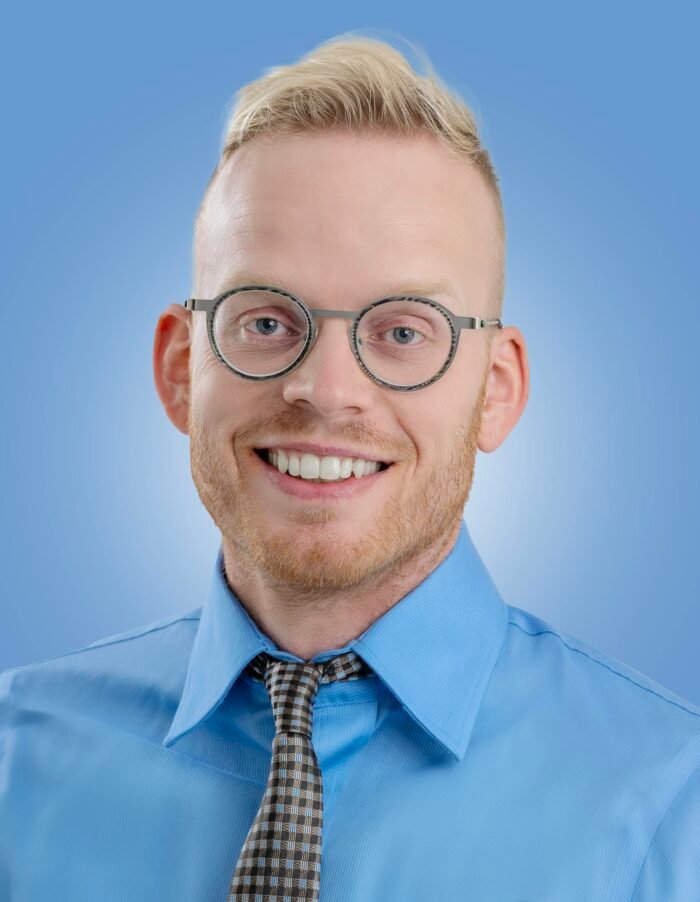 A confident and smiling man in a blue shirt and glasses against a plain background, showcasing a friendly demeanor.