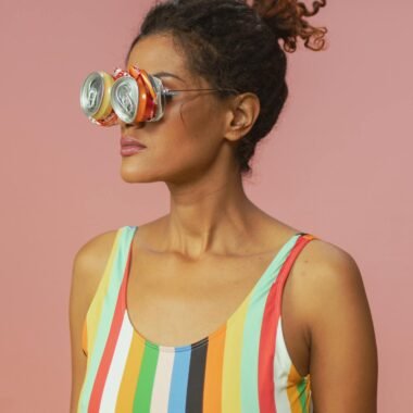Woman in colorful swimsuit wearing recycled can sunglasses against pink background.