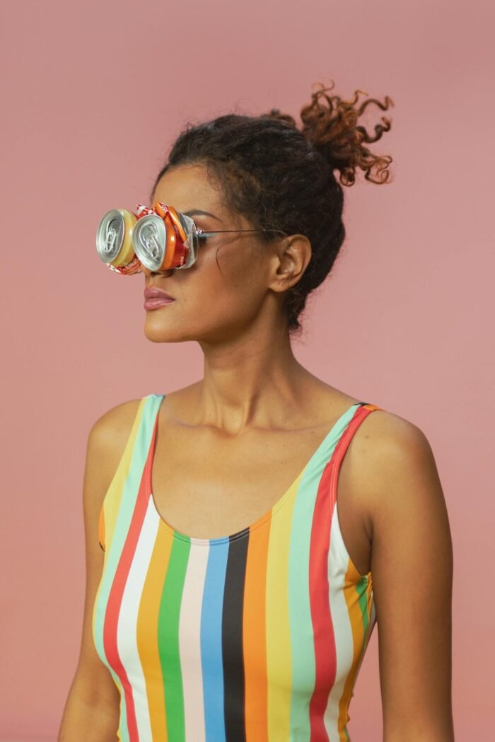 Woman in colorful swimsuit wearing recycled can sunglasses against pink background.